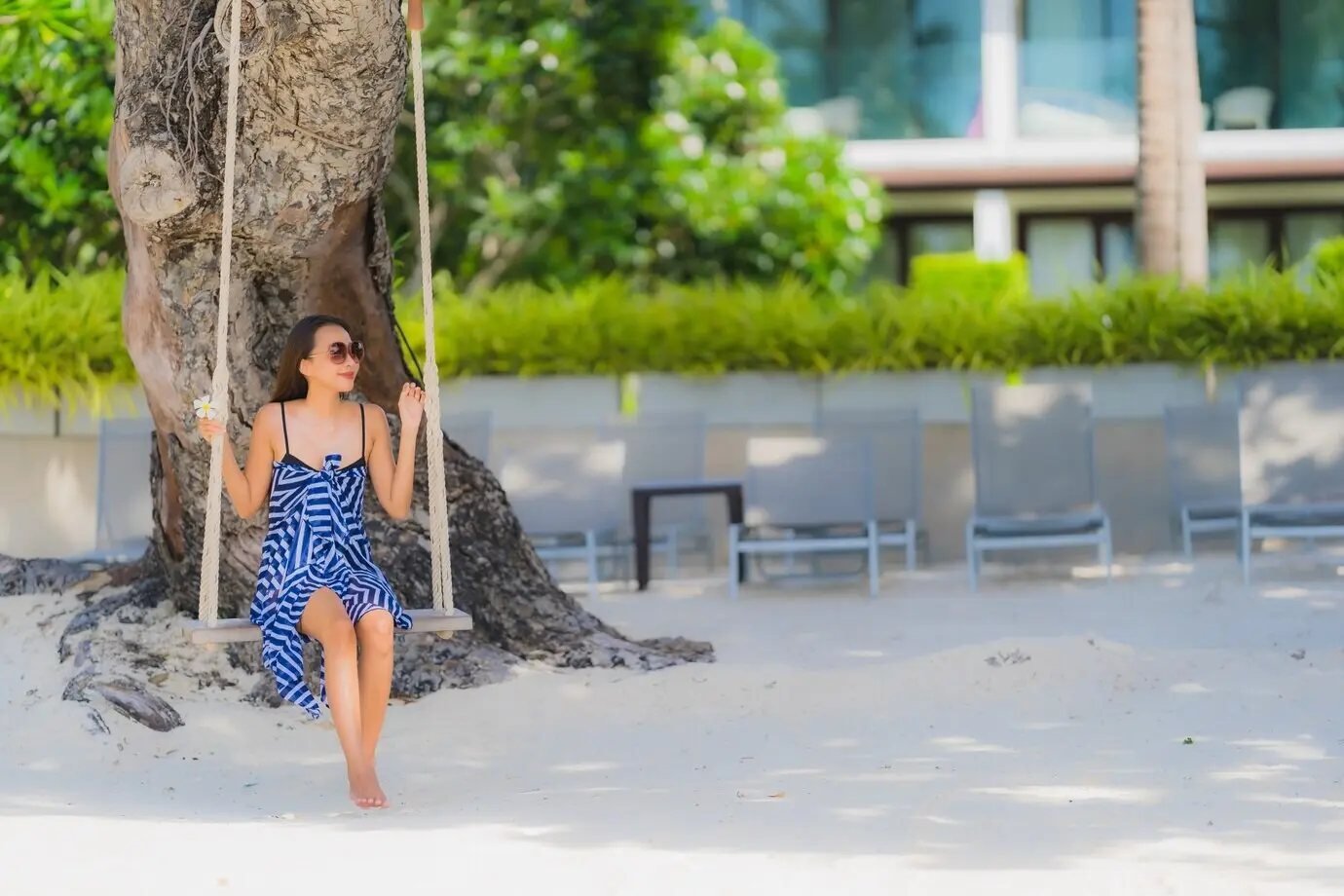 Porträt einer jungen asiatischen Frau, die auf einer Seilschaukel am Strand sitzt, umgeben von Meer und Ozean, mit einer Kokospalme.