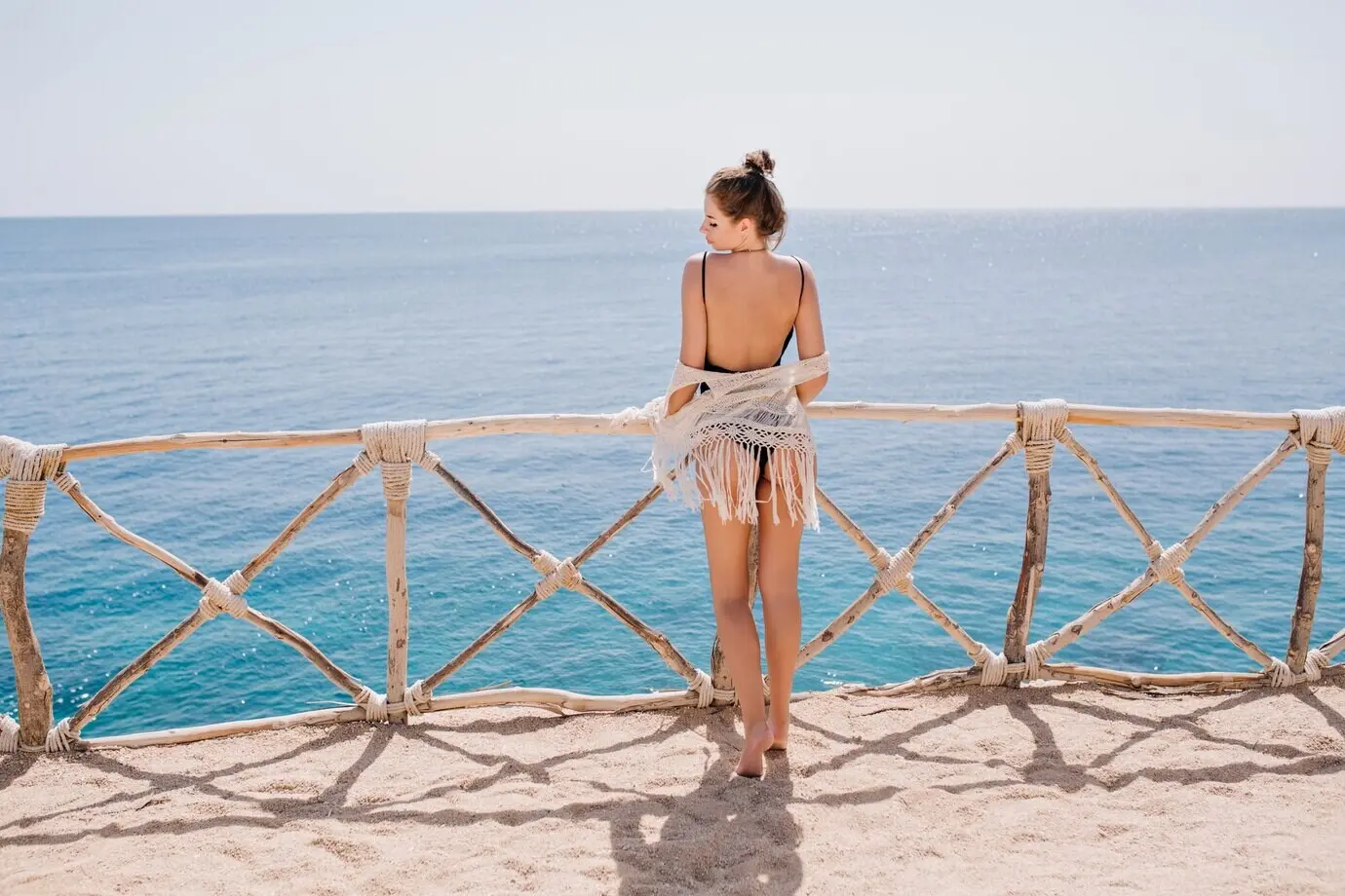 Schlankes, gebräuntes Mädchen mit niedlicher Frisur in Strickkleidung steht auf einer Terrasse und blickt auf das Meer. Beeindruckende junge Frau genießt im Sommerurlaub den Blick auf den Ozean und posiert vor dem Wasser.